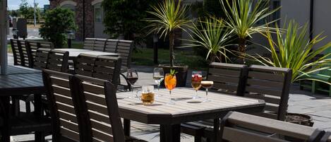 An outdoor patio with wooden tables and chairs, potted plants, and a stone building in the background.
