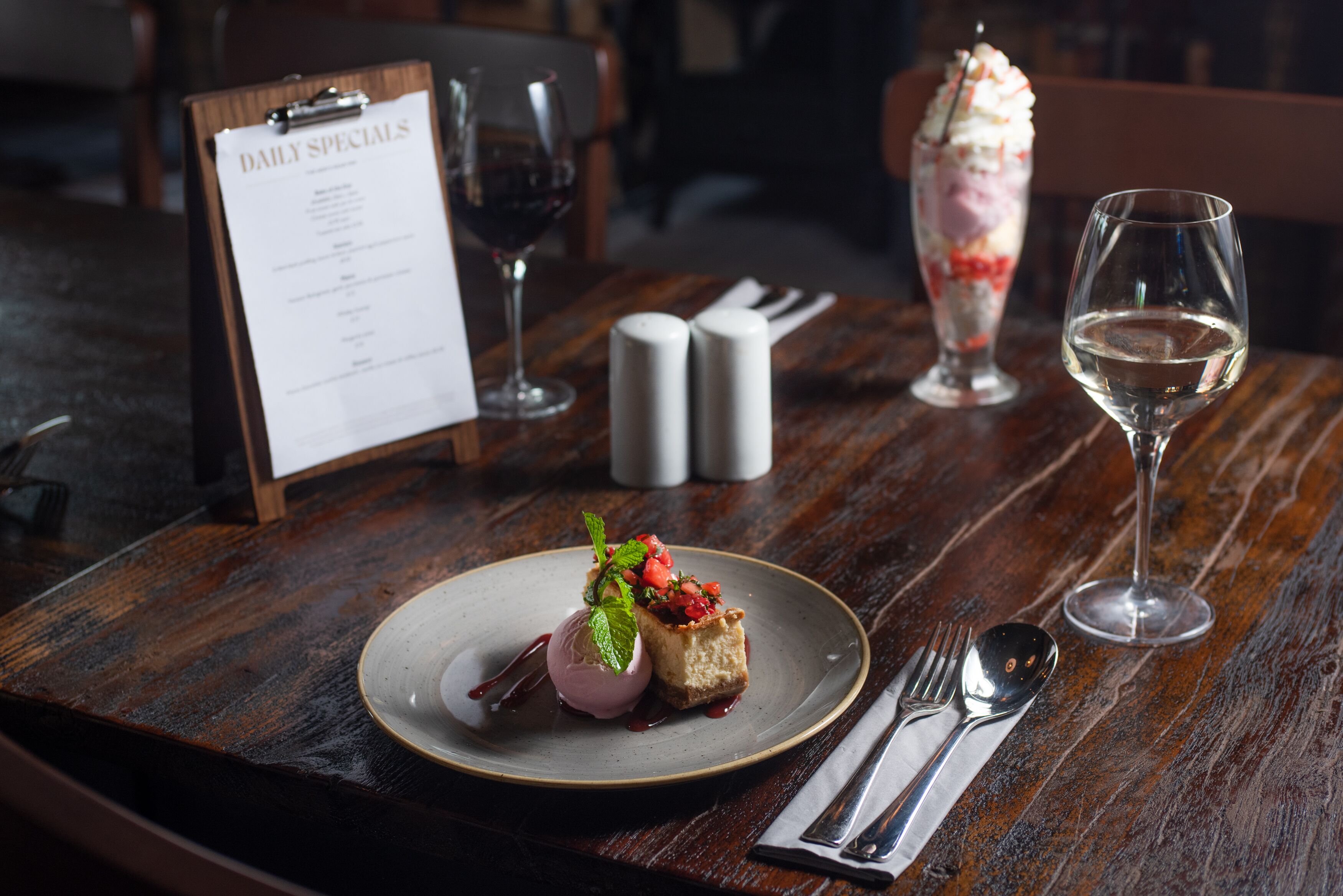 A dessert plate with ice cream and fruit on a wooden table, a menu, salt and pepper shakers, a wine glass, and cutlery.
