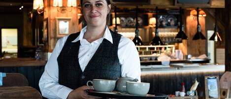 En person holder en bakke med teskeer og et tepot i en restaurant med træindretning og en menutavle.