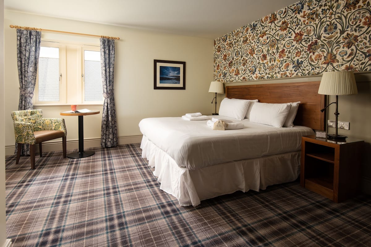 A neatly arranged hotel room with a large bed, a chair, a small table, and a lamp.