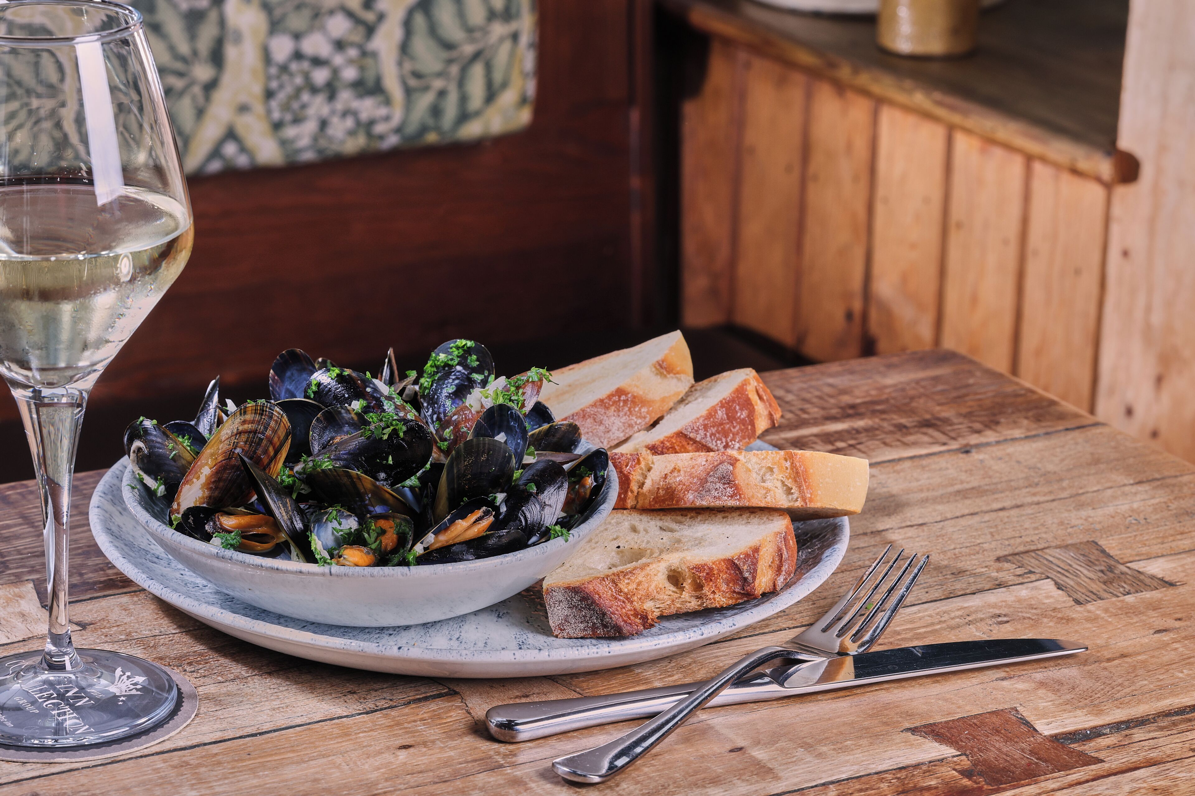 A bowl of mussels with bread on a wooden table, a glass of white wine, and cutlery.