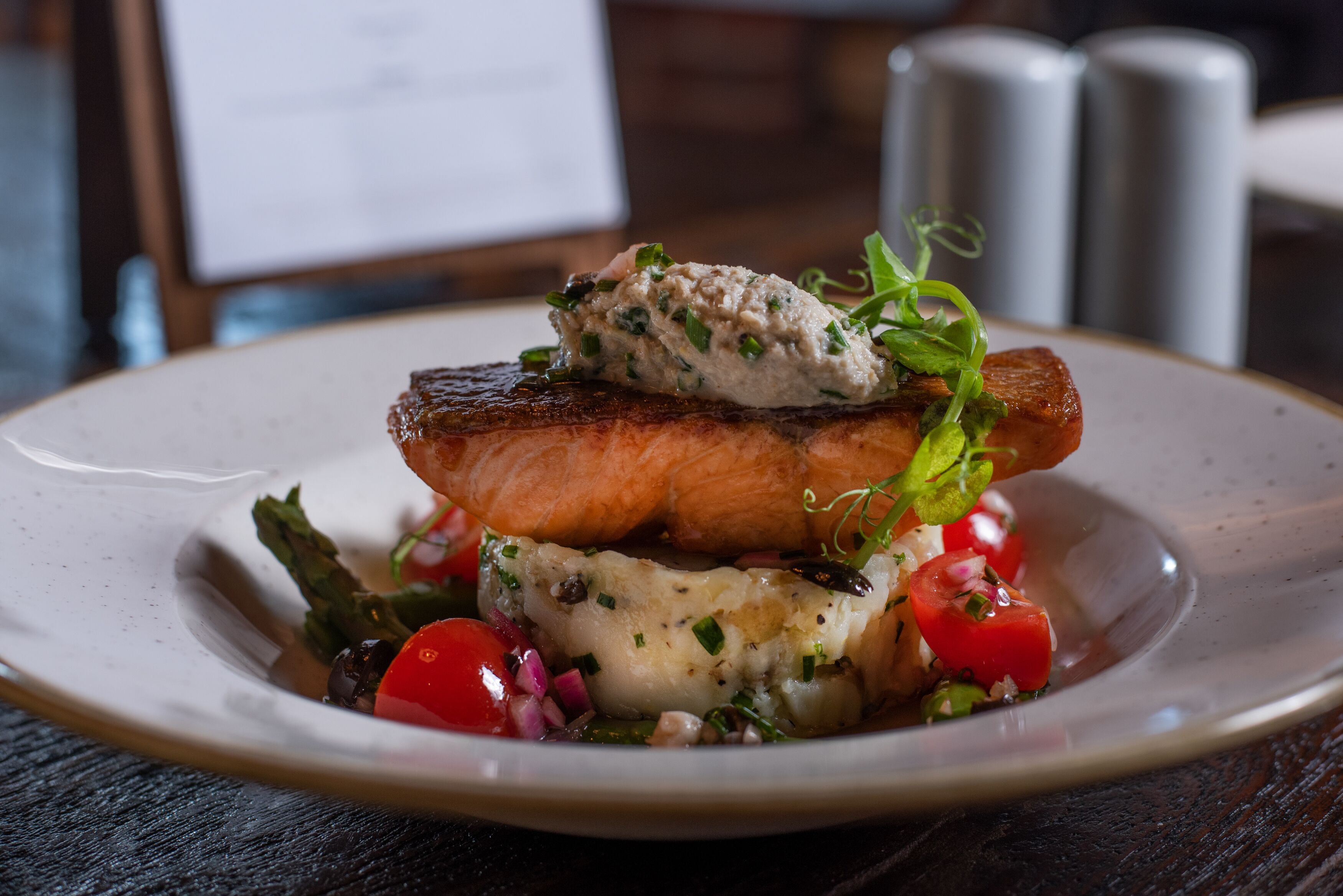 A plate with a grilled salmon fillet, a piece of grilled meat, and a creamy sauce with herbs, accompanied by cherry tomatoes and greens.
