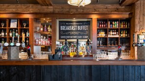 Zona de bar con un letrero de bufé de desayuno, estantes con botellas y un mostrador con vasos y una caja registradora.