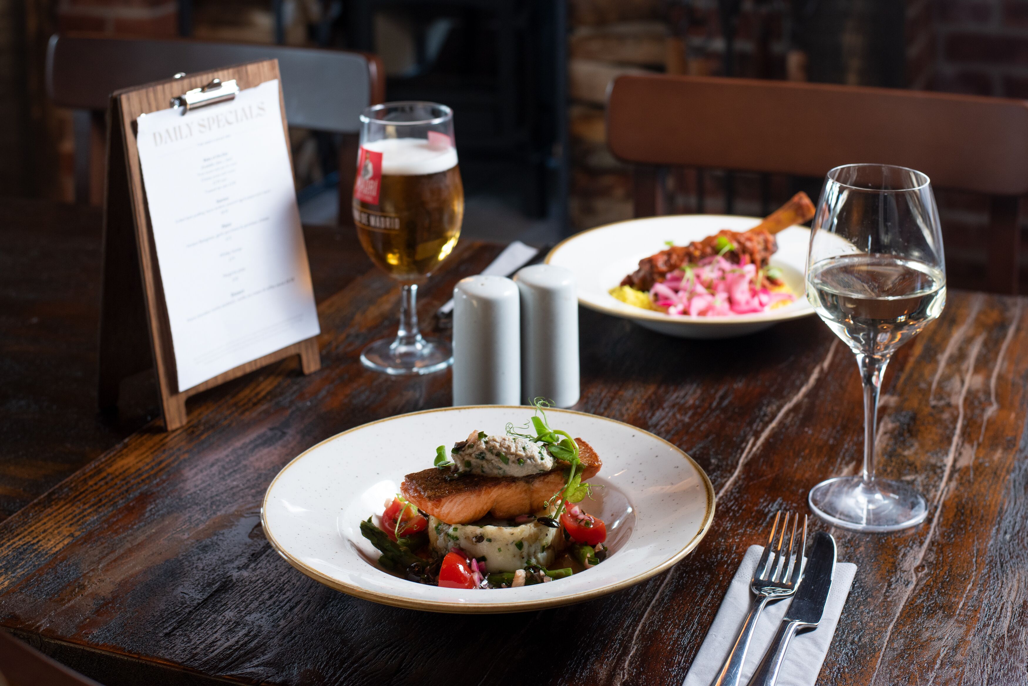 A wooden table with a menu, a glass of beer, a plate of food, and a glass of water.