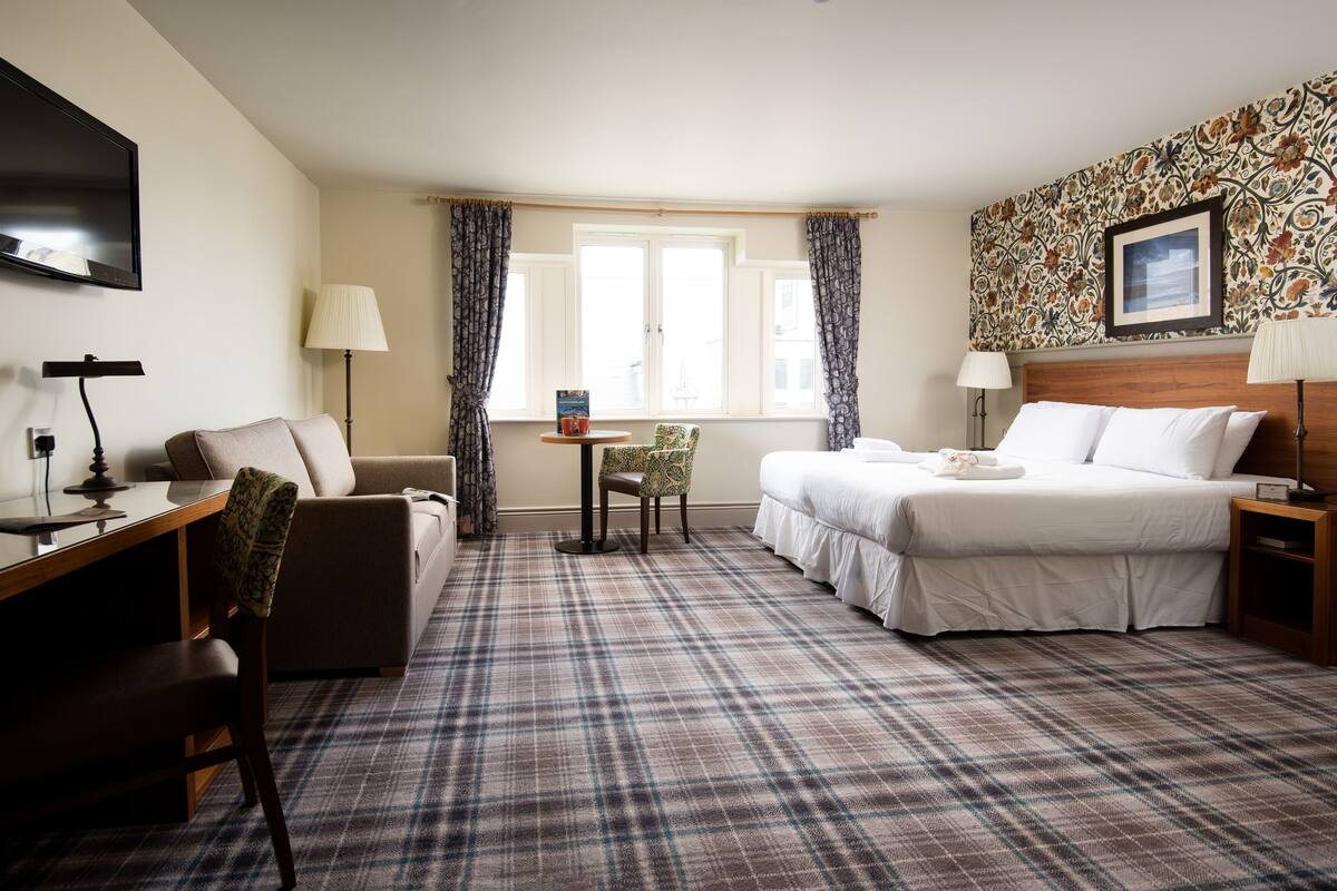 A hotel room with a bed, a desk, a chair, a small table, and a TV.