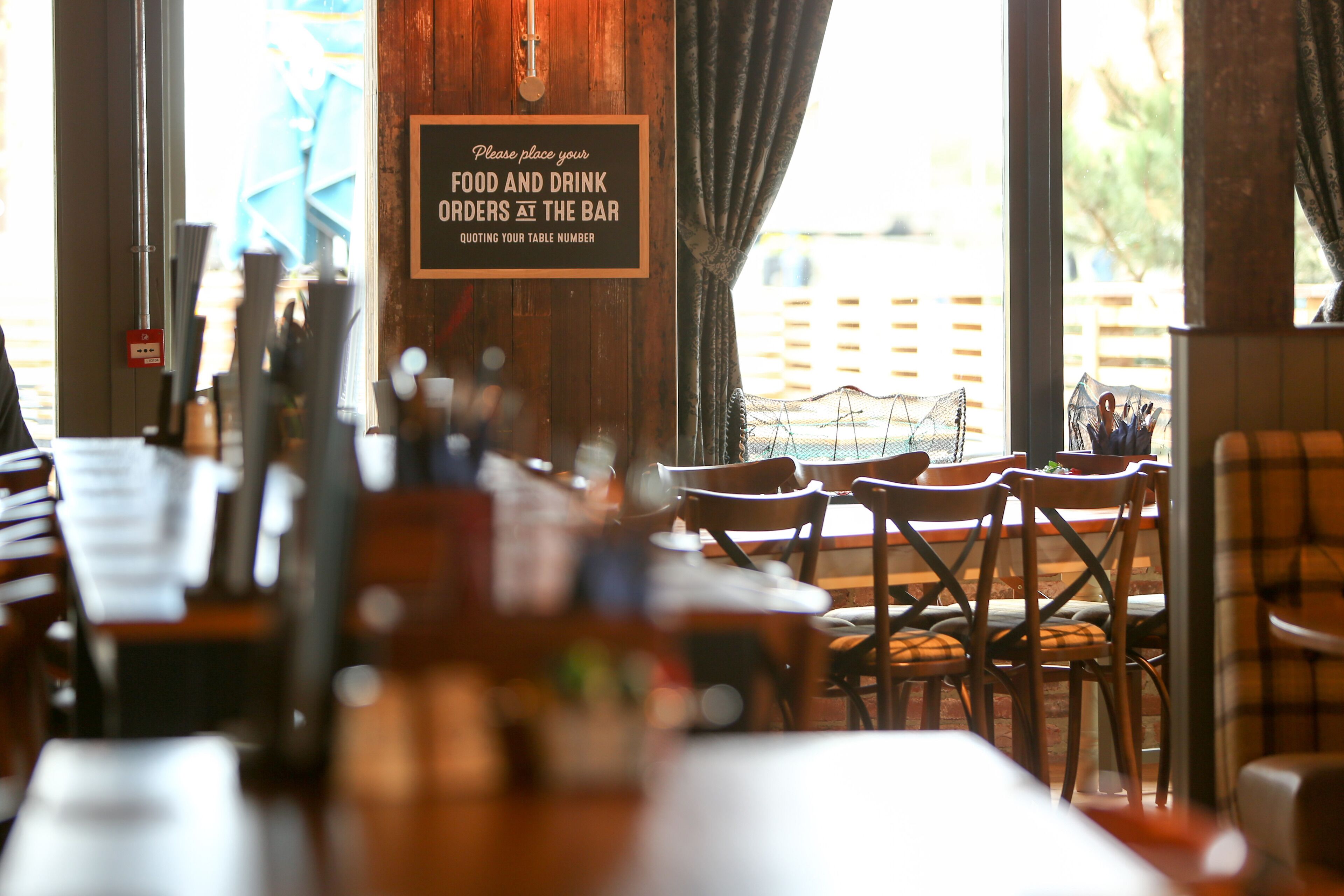 A restaurant interior with wooden tables and chairs, a sign for food and drink orders at the bar, and a view of a wooden deck outside.