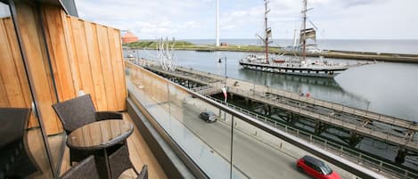 A balcony with wicker chairs, a view of a river with a sailing ship and wind turbines, and a road with a red car.