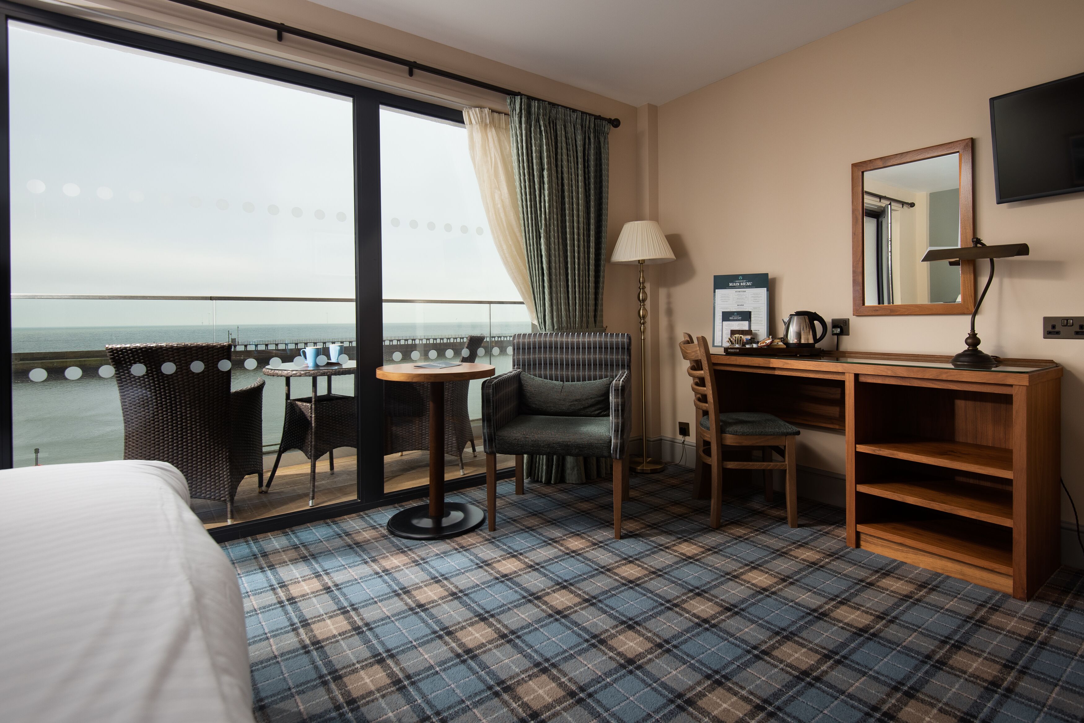 A hotel room with a bed, a desk, a chair, a small table, a lamp, a mirror, and a view of the sea through a large window.
