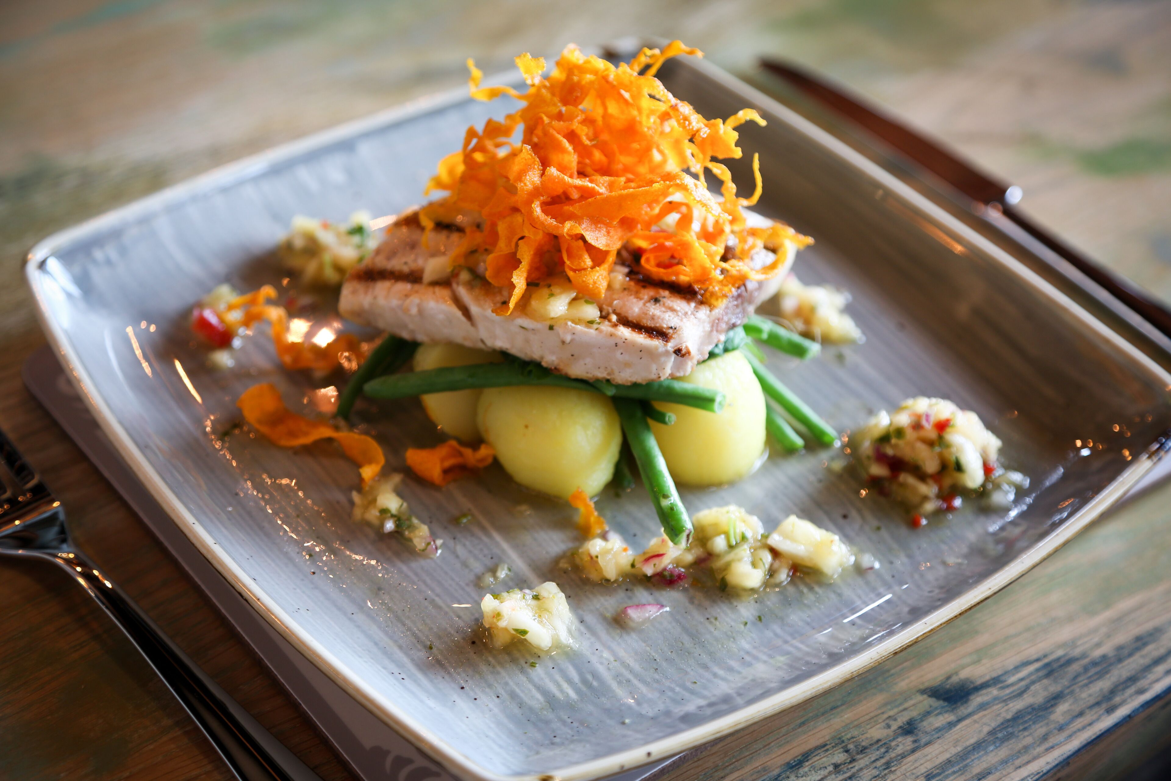 A square plate with a grilled fish topped with shredded orange vegetable, green beans, and a side of mashed potatoes.