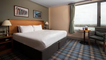 Habitación de hotel con una cama grande, dos mesitas de noche con lámparas, una pequeña mesa redonda con una silla y vistas al mar a través de una ventana.