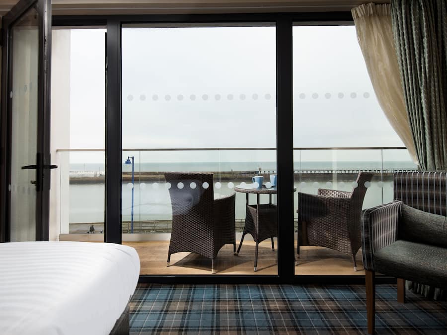 A hotel room with a large sliding glass door leading to a balcony with wicker furniture and a view of the sea.