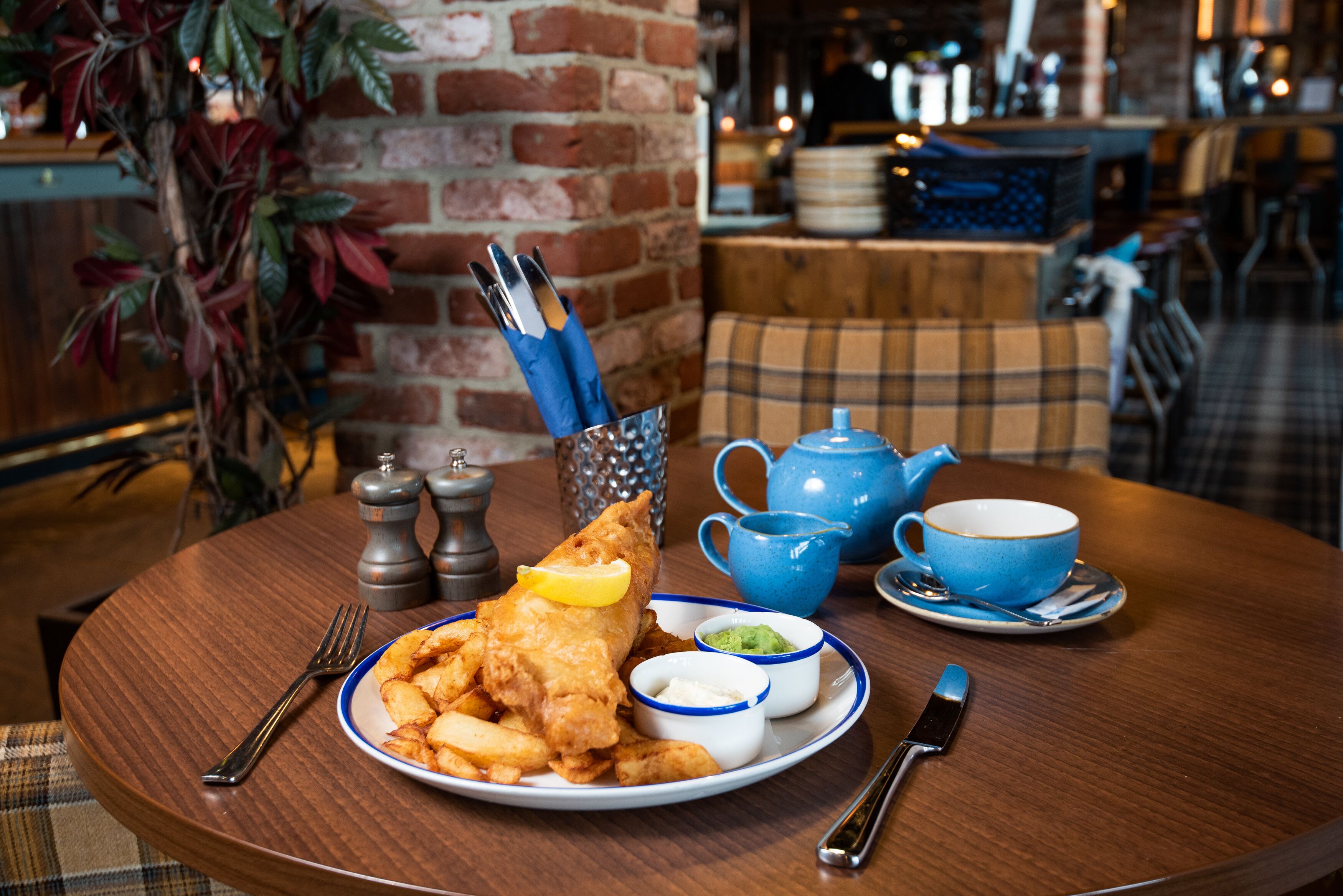 A table with a plate of fish and chips, a teapot, cups, and condiments.