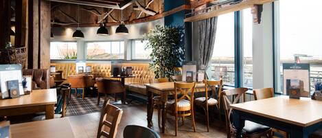A cozy restaurant interior with wooden furniture, a large plant, and a view through the windows.