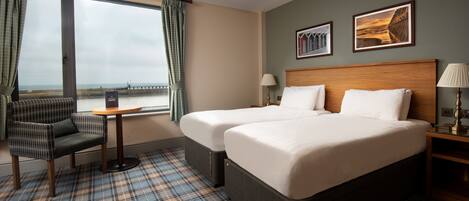 A hotel room with two beds, a chair, a small table, and a view of the sea through large windows.