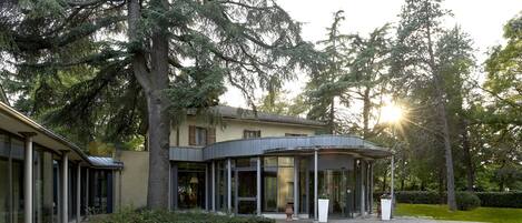 A modern building with a curved glass entrance, surrounded by lush greenery and large trees.