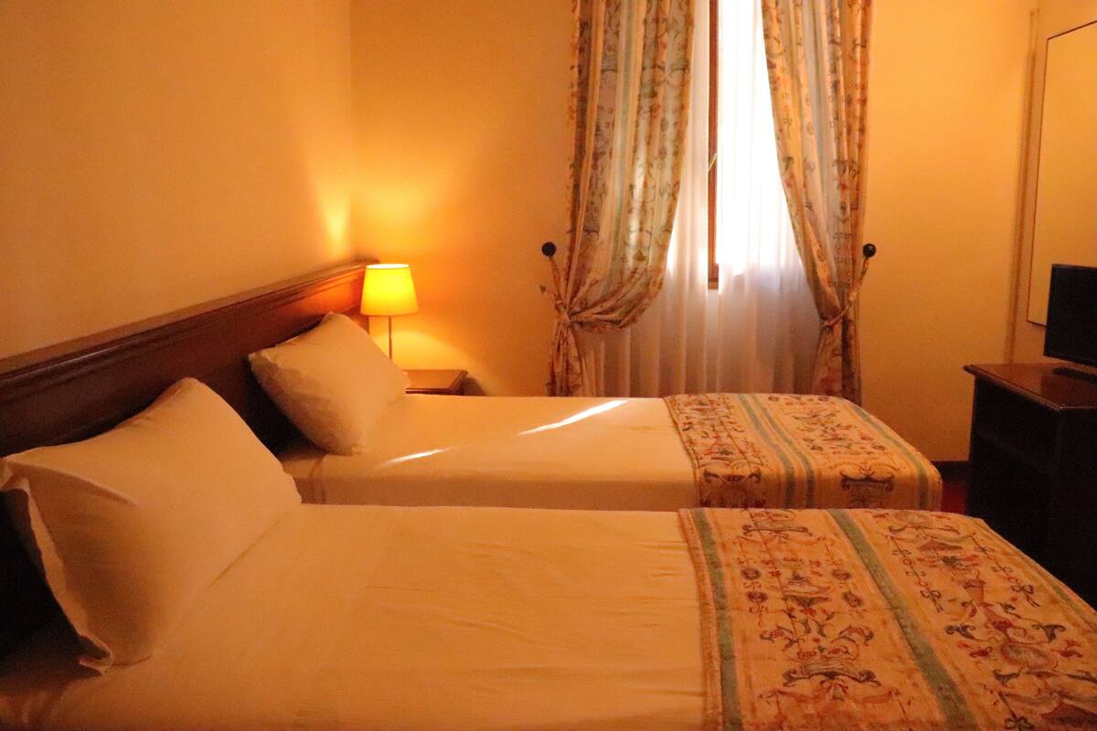 A hotel room with two beds, a nightstand with a lamp, a TV on a stand, and floral curtains.