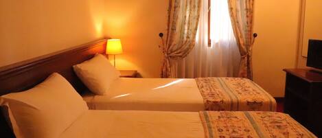 Una habitación de hotel con dos camas, una mesita de noche con lámpara, un televisor en un mueble bajo y cortinas con estampado floral.