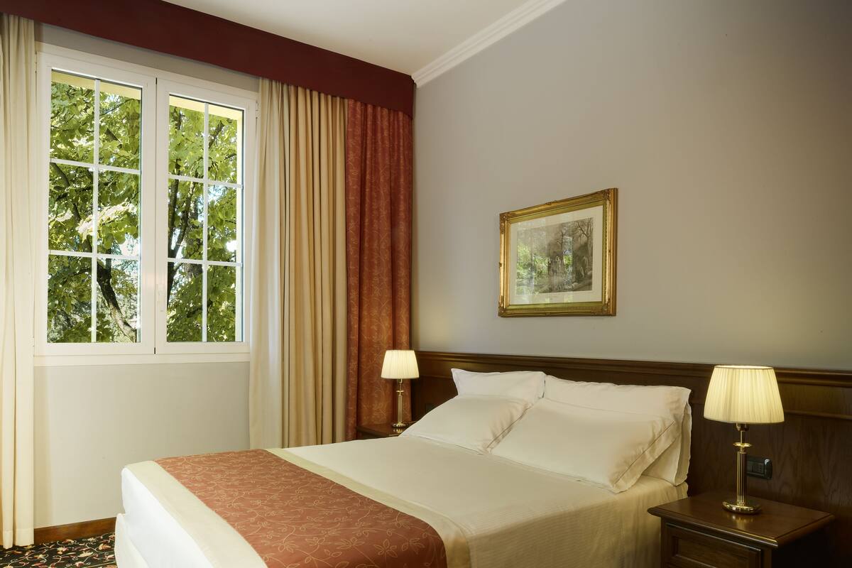 A hotel room with a large bed, two bedside tables with lamps, a window with curtains, and a framed picture on the wall.