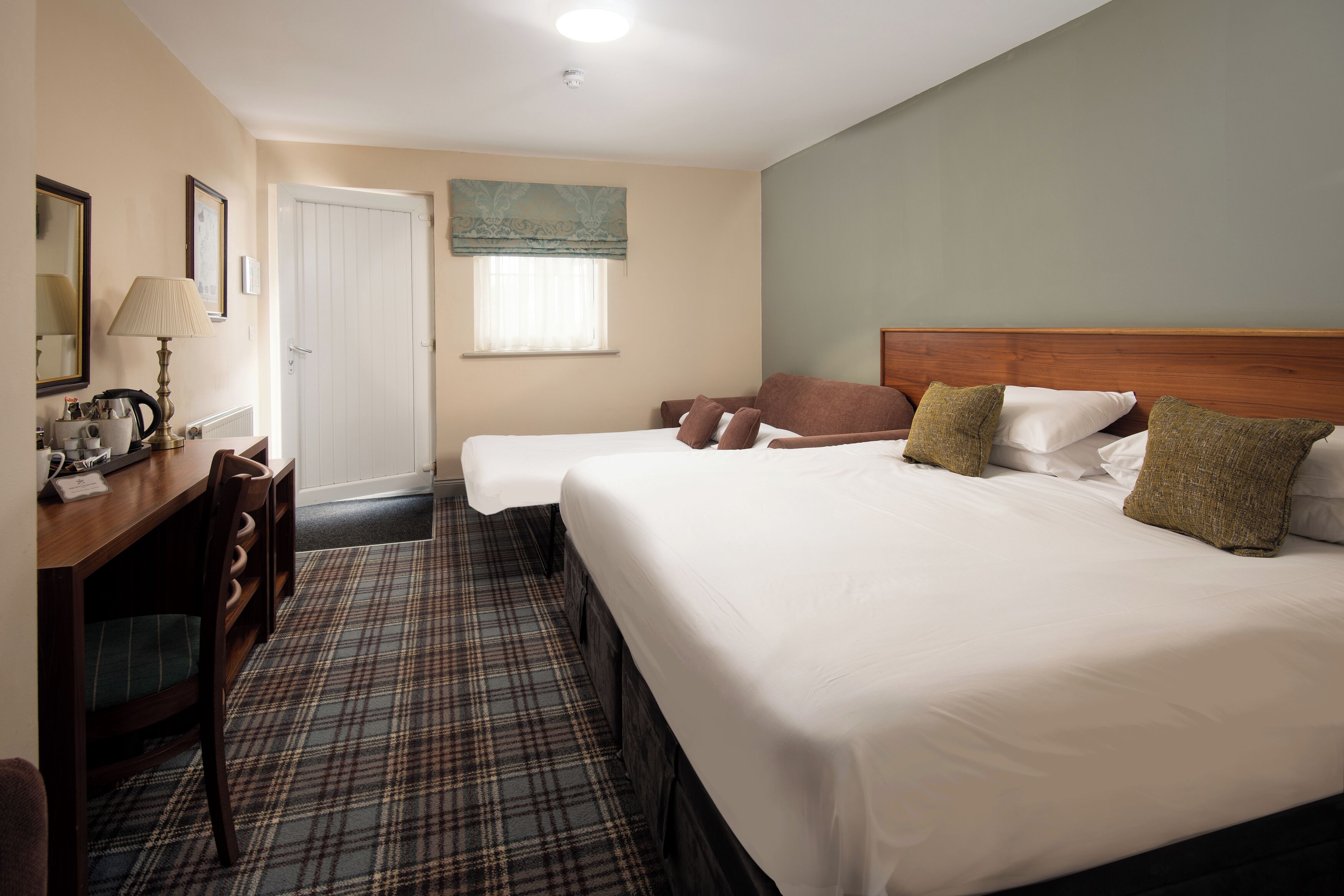 A hotel room with two beds, a desk with a lamp, a chair, a window with curtains, and a door.