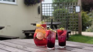 A wooden table with a pitcher and two glasses of iced drinks, a wooden fence, and a hanging flower basket in the background. - The Lindisfarne Inn - The Inn Collection Group (Berwick-upon-Tweed)