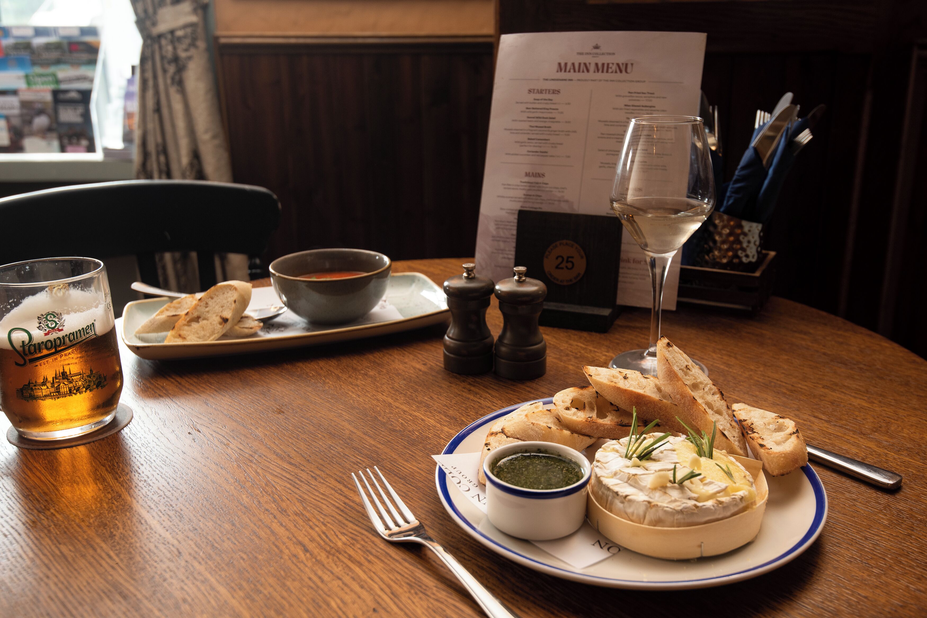 Een eettafel met een bord kaas, brood en een glas bier. Een menukaart is op de achtergrond zichtbaar.