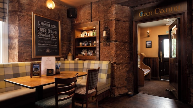 Un restaurant accueillant avec un mur en pierre, une table en bois entourée de chaises, un banc à carreaux et un tableau affichant le menu, ainsi que des offres spéciales pour les chambres avec salle de bain privée et les formules de petit-déjeuner.