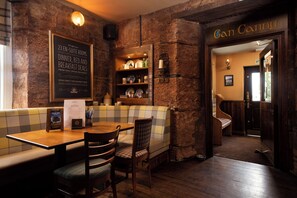 A cozy restaurant with a stone wall, a wooden table with chairs, a checkered bench, and a menu board advertising en-suite rooms and breakfast deals. - The Lindisfarne Inn - The Inn Collection Group (Berwick-upon-Tweed)