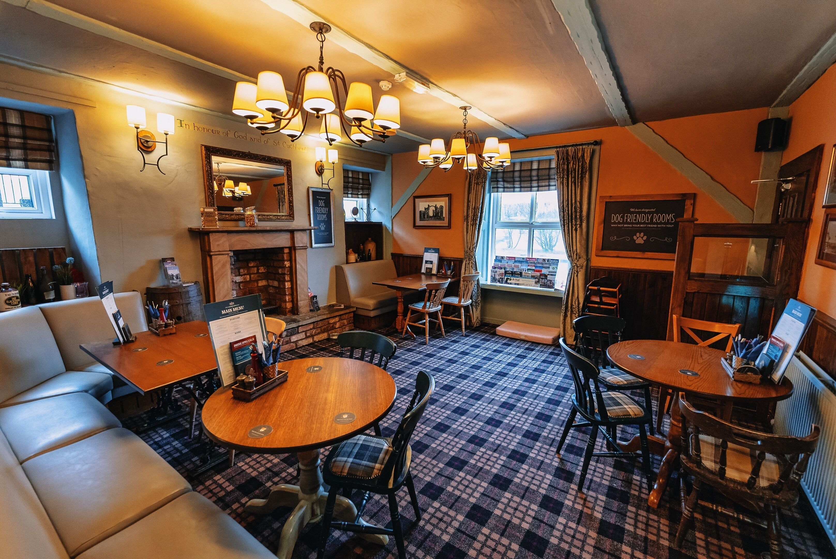 A cozy pub with a checkered floor, wooden furniture, and a fireplace. There are tables and chairs for dining, and a sign indicating dog-friendly rooms.