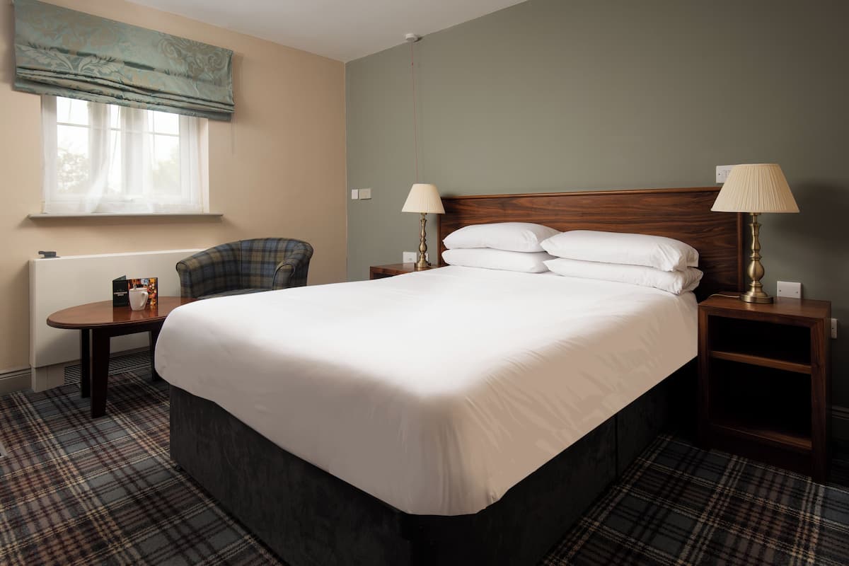 Une chambre d’hôtel avec un grand lit, deux tables de chevet avec des lampes, une chaise, une fenêtre avec des rideaux et un radiateur.