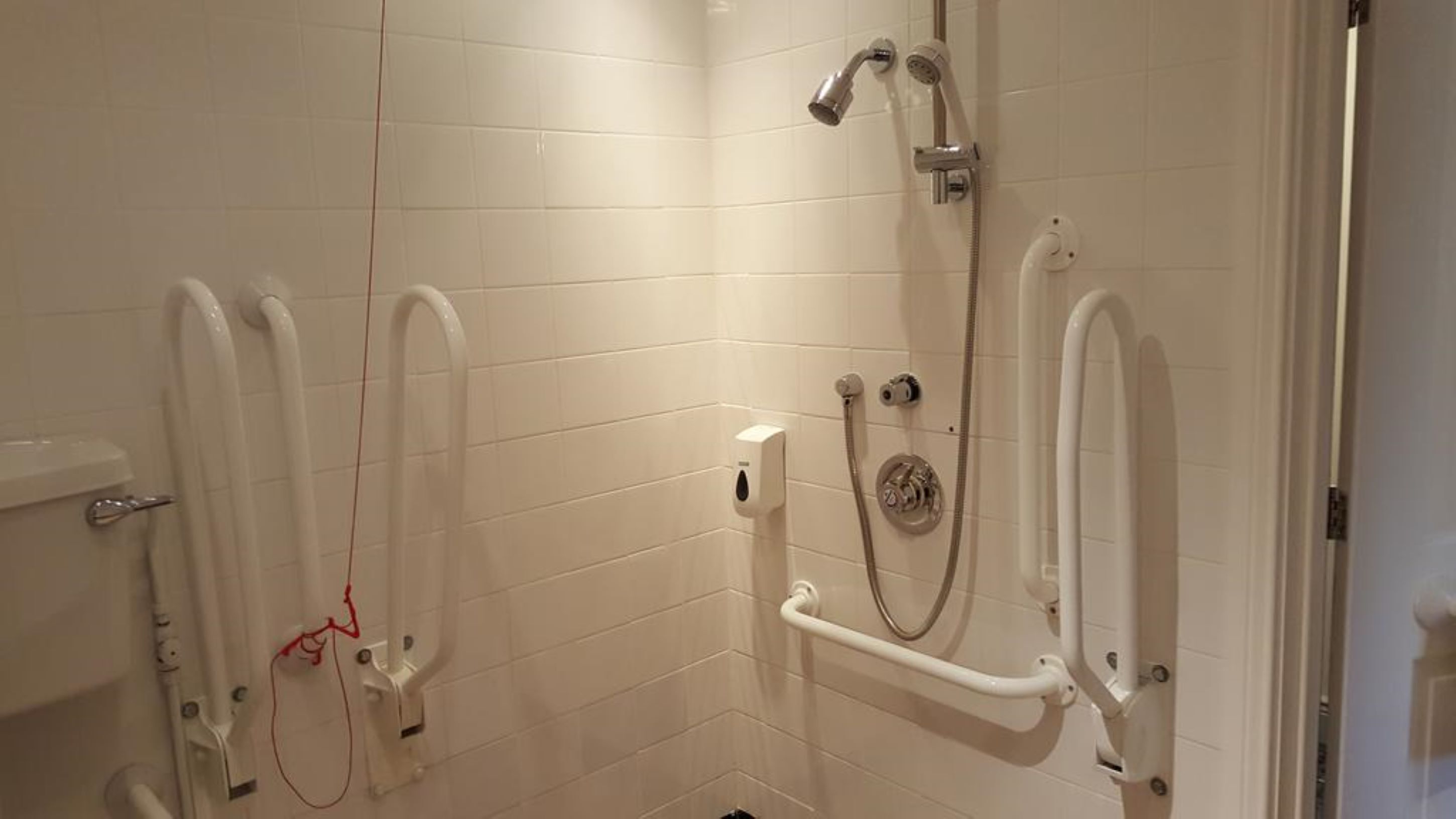 A bathroom with white tiled walls, a shower with a handheld showerhead, and a grab bar.