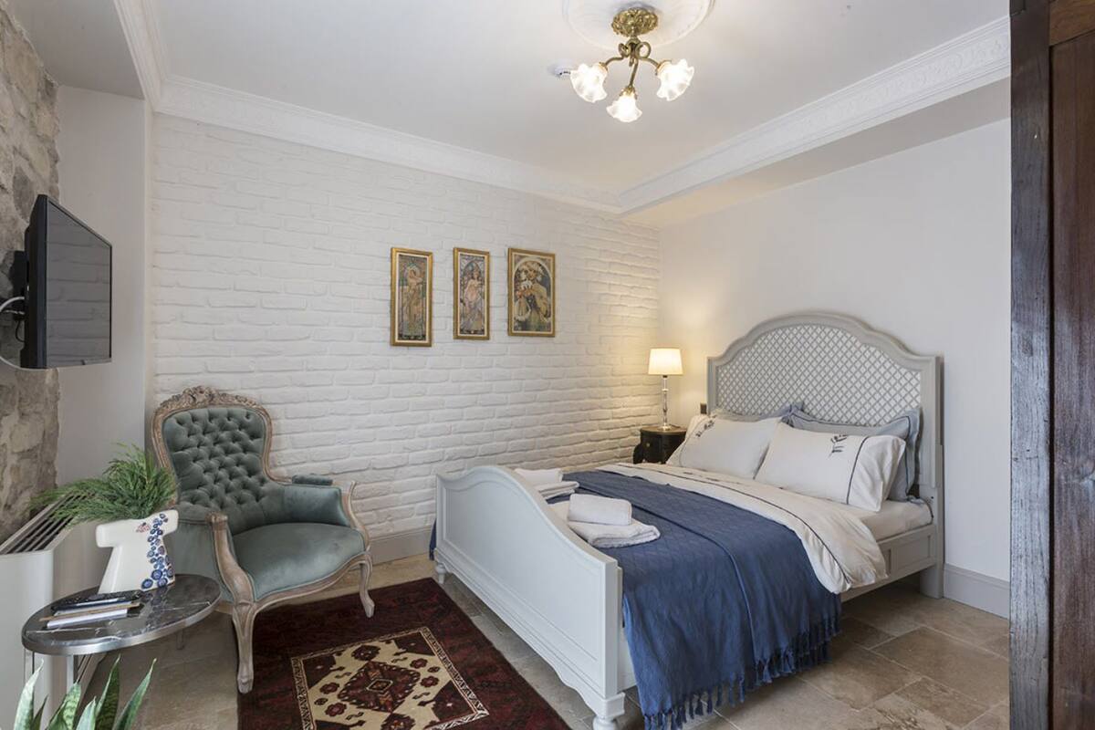 Une chambre avec un mur en briques blanches, un lit recouvert d’une couverture bleue, un fauteuil gris et une petite table sur laquelle se trouvent une plante et une lampe.