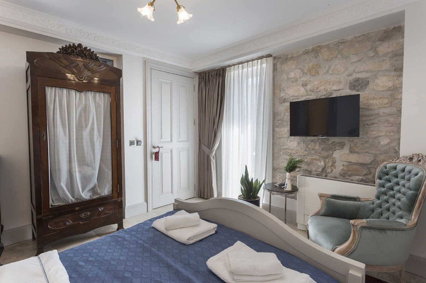 A bedroom with a bed, a vanity with a mirror, a stone wall, a television, and a chair.