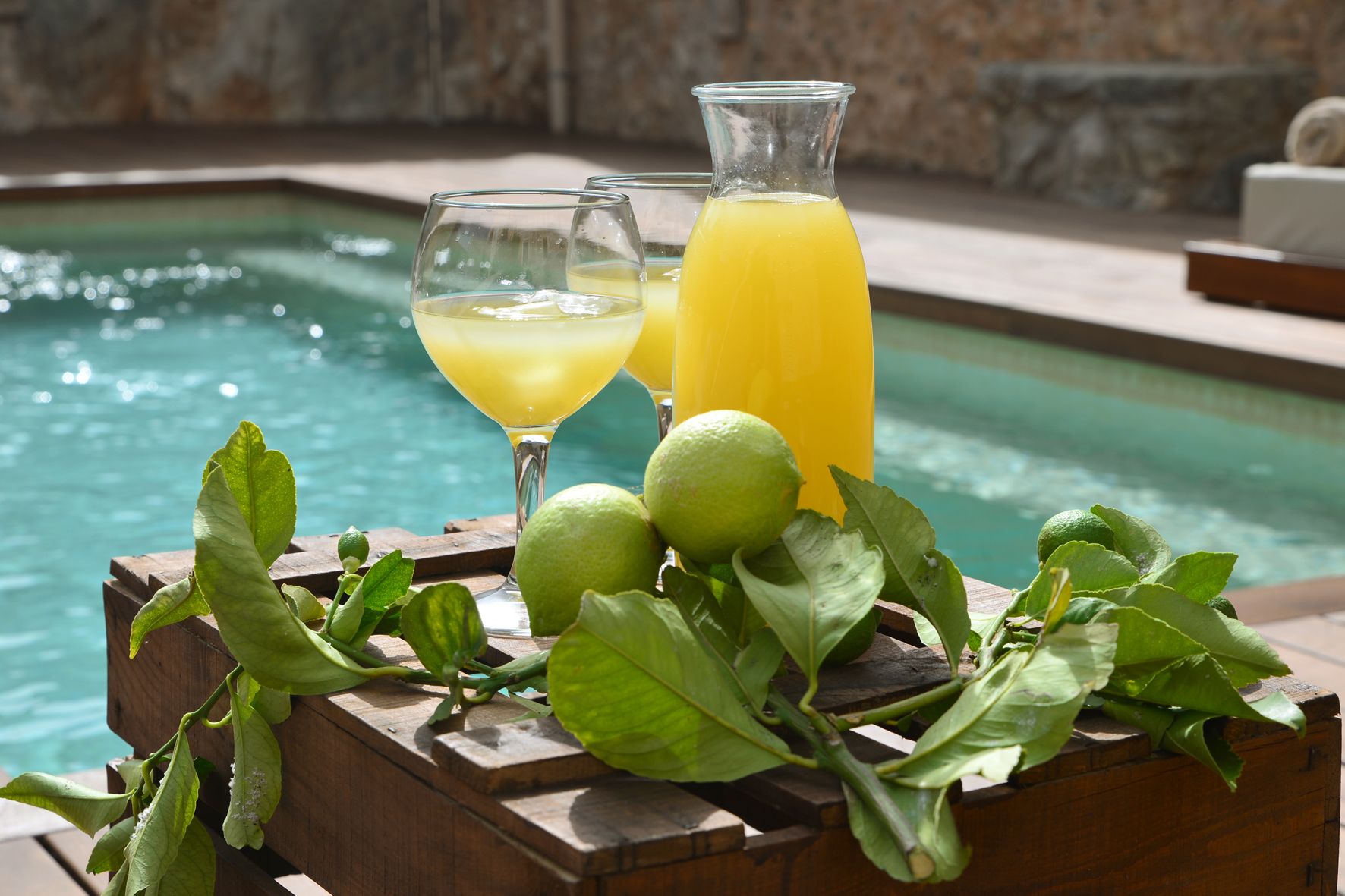 A wooden table with a glass of orange juice, a glass of white wine, and a bottle of orange juice by a pool.