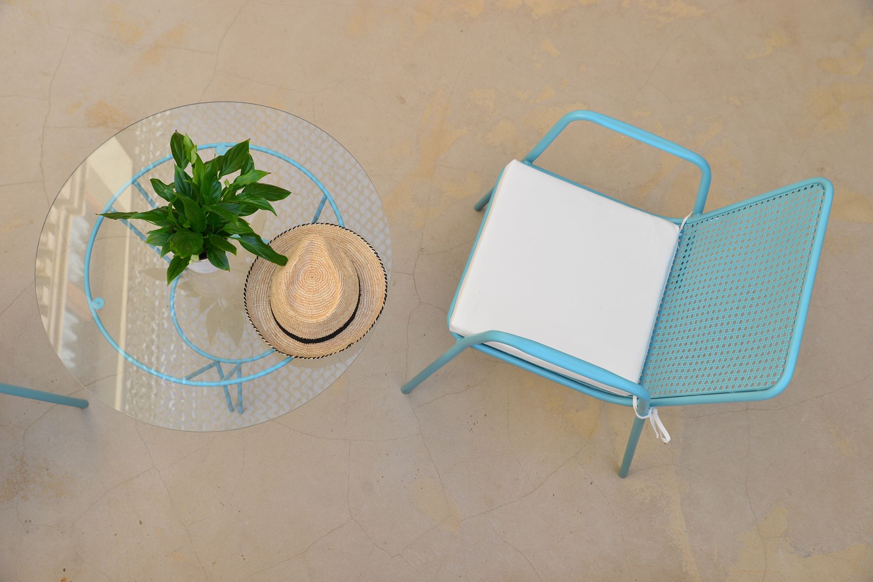 A round glass table with a potted plant and a straw hat, and a blue metal chair with a white cushion.