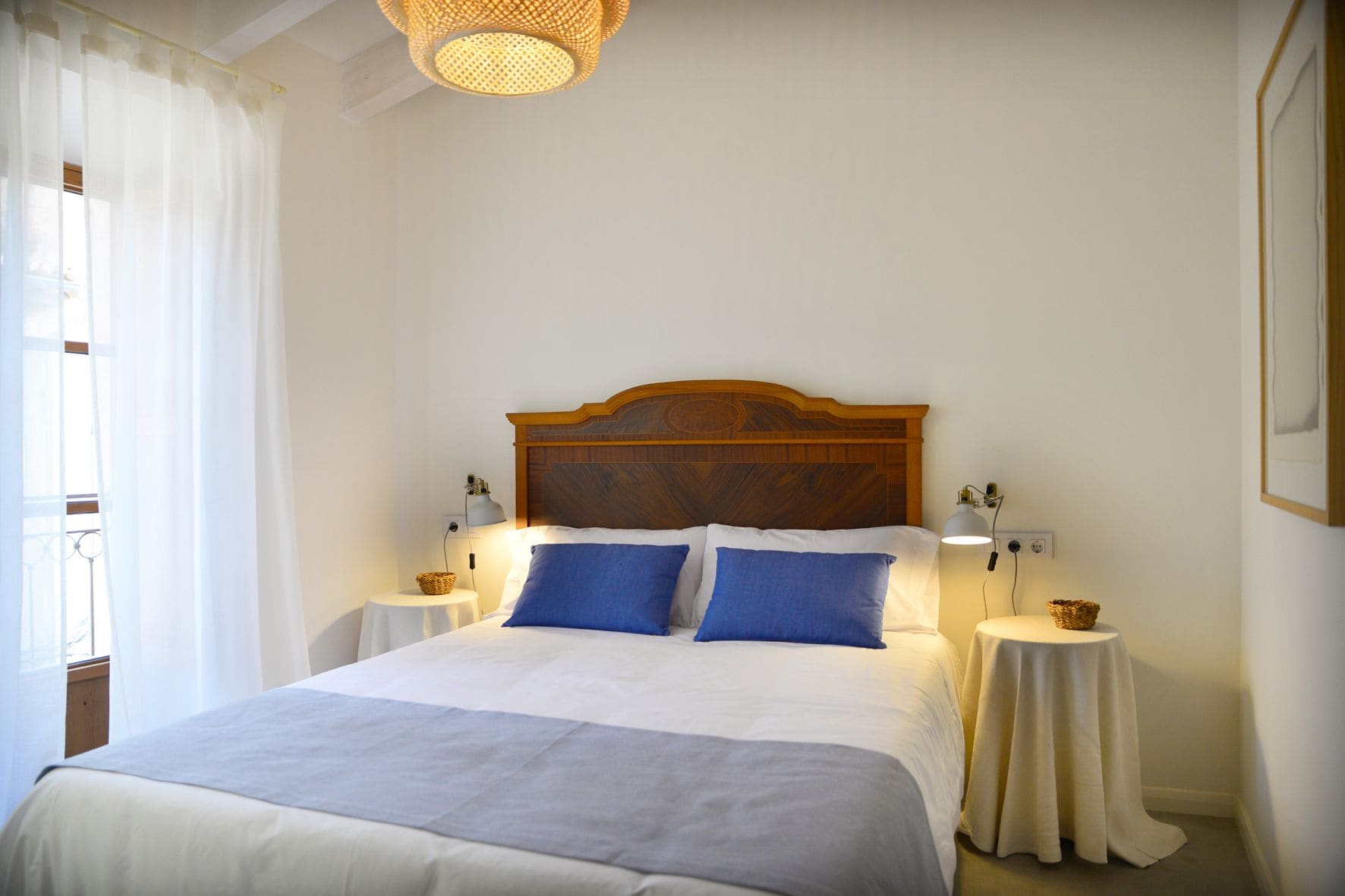 A neatly made bed with a wooden headboard, two bedside tables with lamps, and a window with sheer curtains.