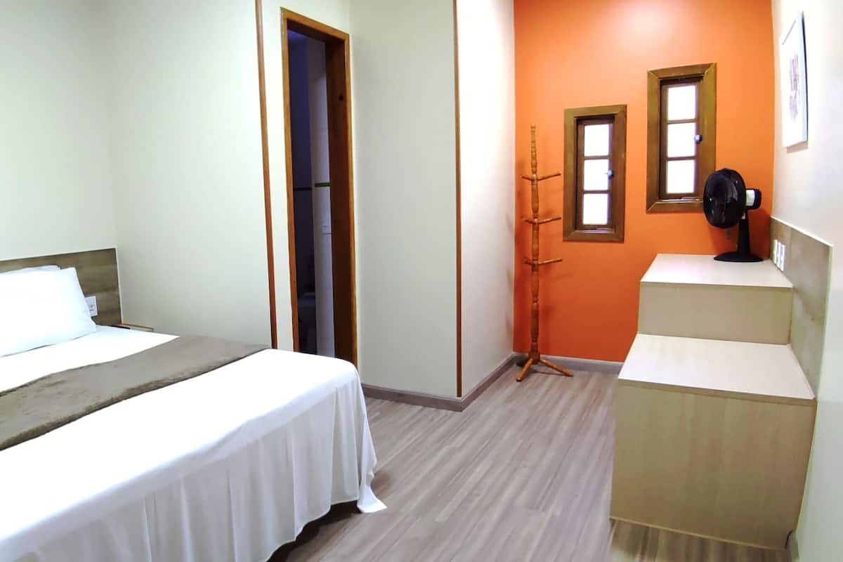 Ein Schlafzimmer mit einem Bett, einem Nachttisch, einem Standventilator und einer Tür zu einem weiteren Raum.
