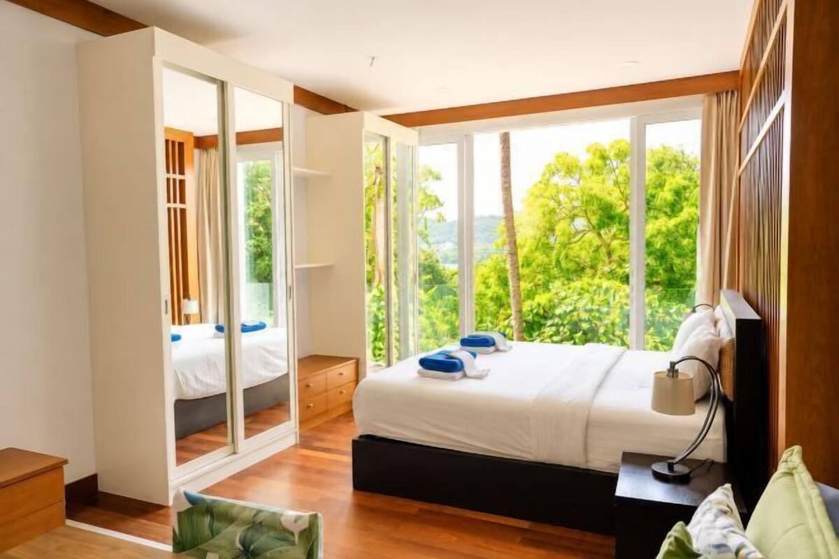 A bedroom with a bed, a nightstand, a mirror, and a window with a view of greenery.