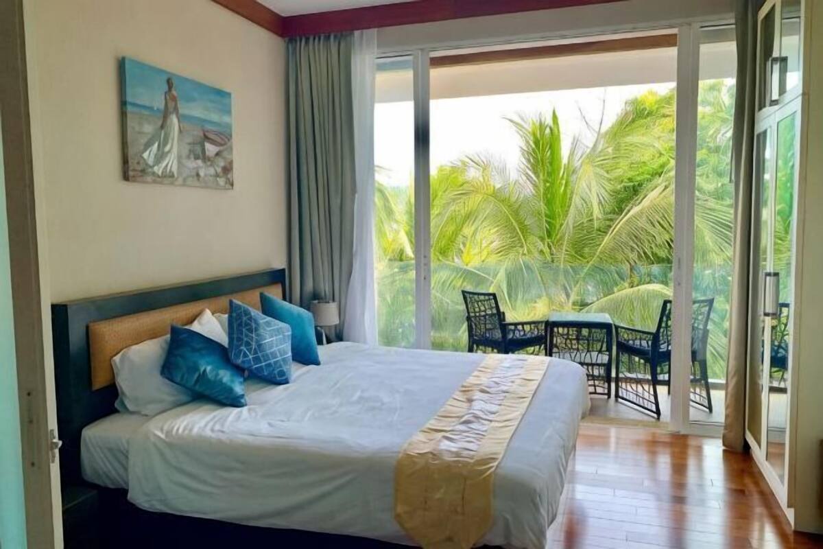 A bedroom with a bed, a painting, a balcony with chairs, and a view of palm trees.