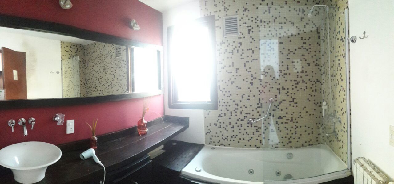 Une salle de bain avec un lavabo blanc, un miroir, une douche dont le mur est recouvert de mosaïques, et une fenêtre laissant entrer la lumière naturelle.