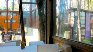 A dining area with white chairs and tables, large windows with a view of trees and buildings. - Altos Medanos - Cabañas y Club de Bosque (Mar de Las Pampas)