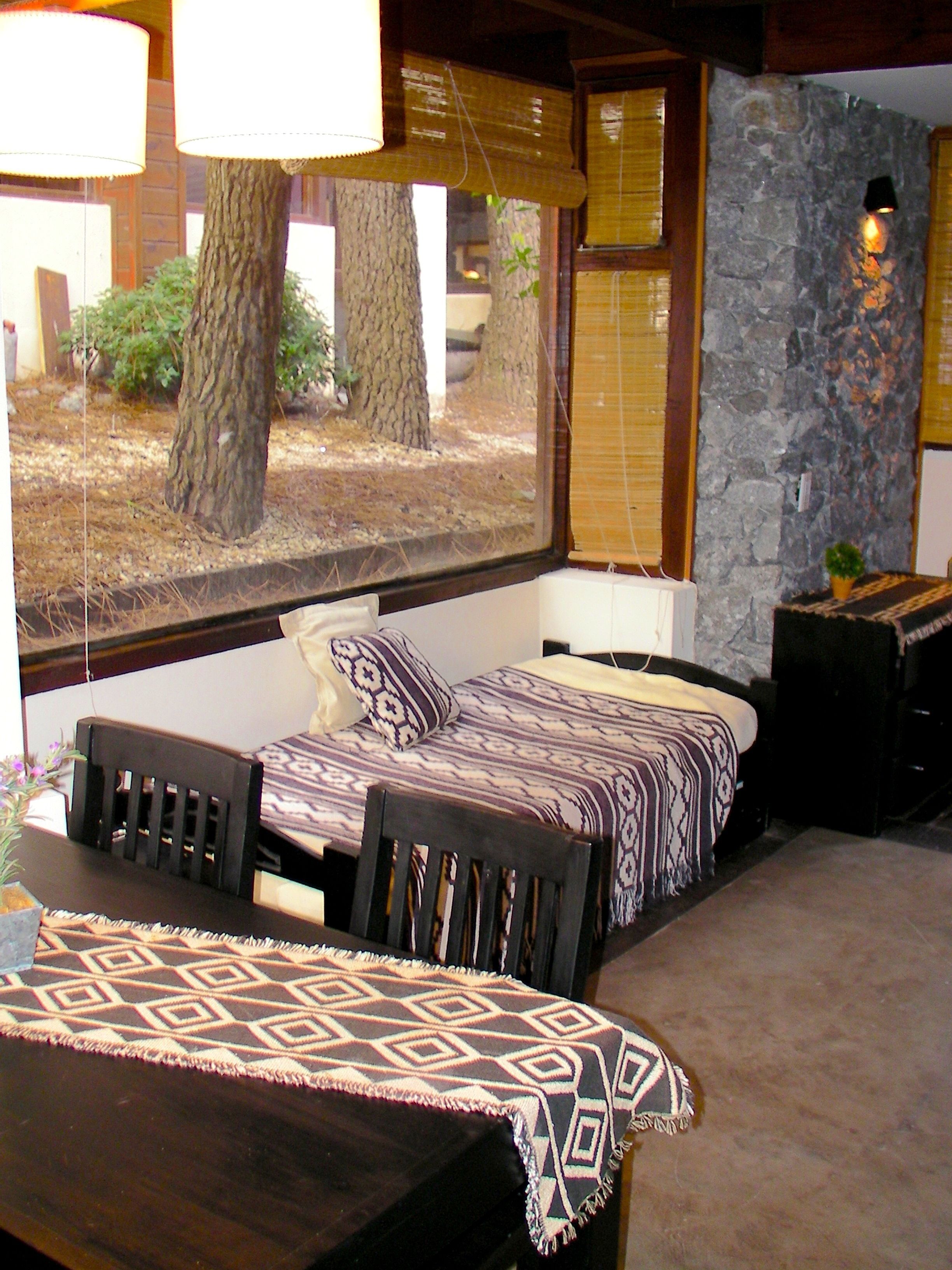 Quarto com cama, janela com vista para árvores, parede de pedra e pequena mesa com toalha de mesa estampada.