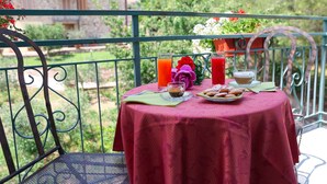 Une table sur un balcon, recouverte d’une nappe rouge, avec deux verres de jus d’orange, une tasse de café, une assiette de pâtisseries et un vase rempli de fleurs.