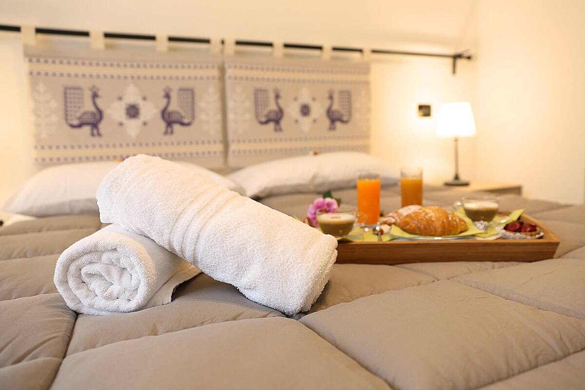 A neatly made bed with a rolled white towel, a tray with croissants, juice, and coffee, and a lamp in a hotel room.