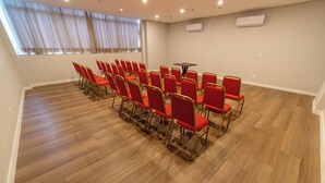 Uma sala de conferências com fileiras de cadeiras vermelhas, piso de madeira e uma grande janela com cortinas.