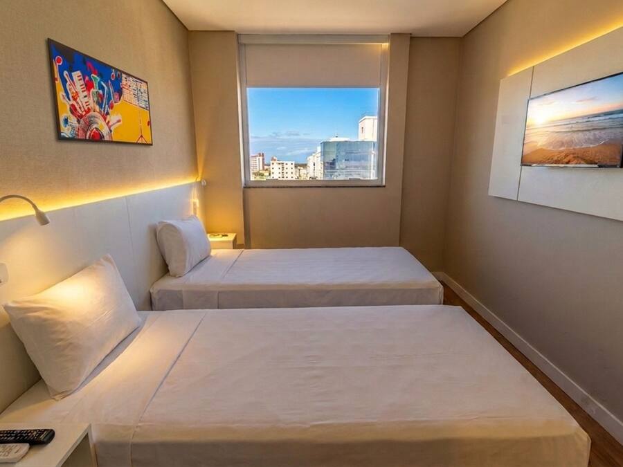 Quarto de hotel com duas camas, uma mesa de cabeceira, um abajur, um quadro e uma janela com vista para a cidade.