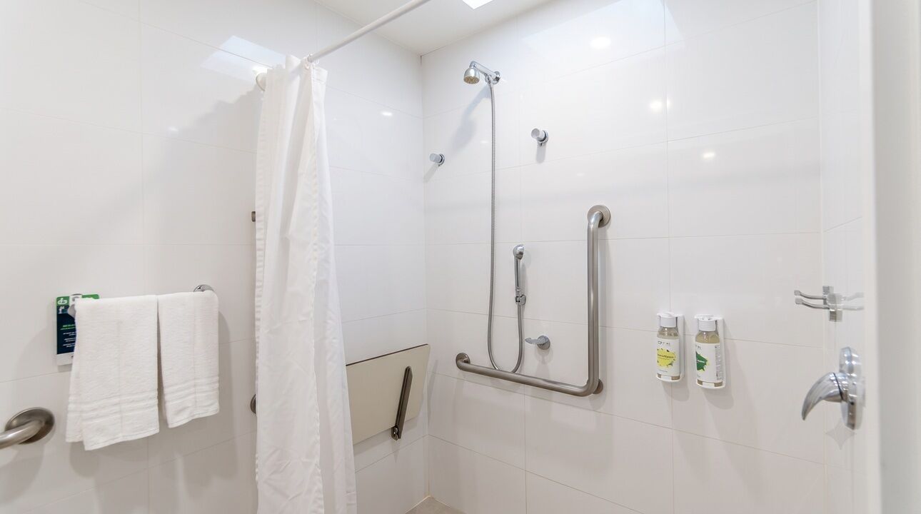 Ein modernes Badezimmer mit weißen Fliesen, einer Dusche inklusive Handbrause, einer Haltestange und einem Handtuchhalter mit zwei Handtüchern.