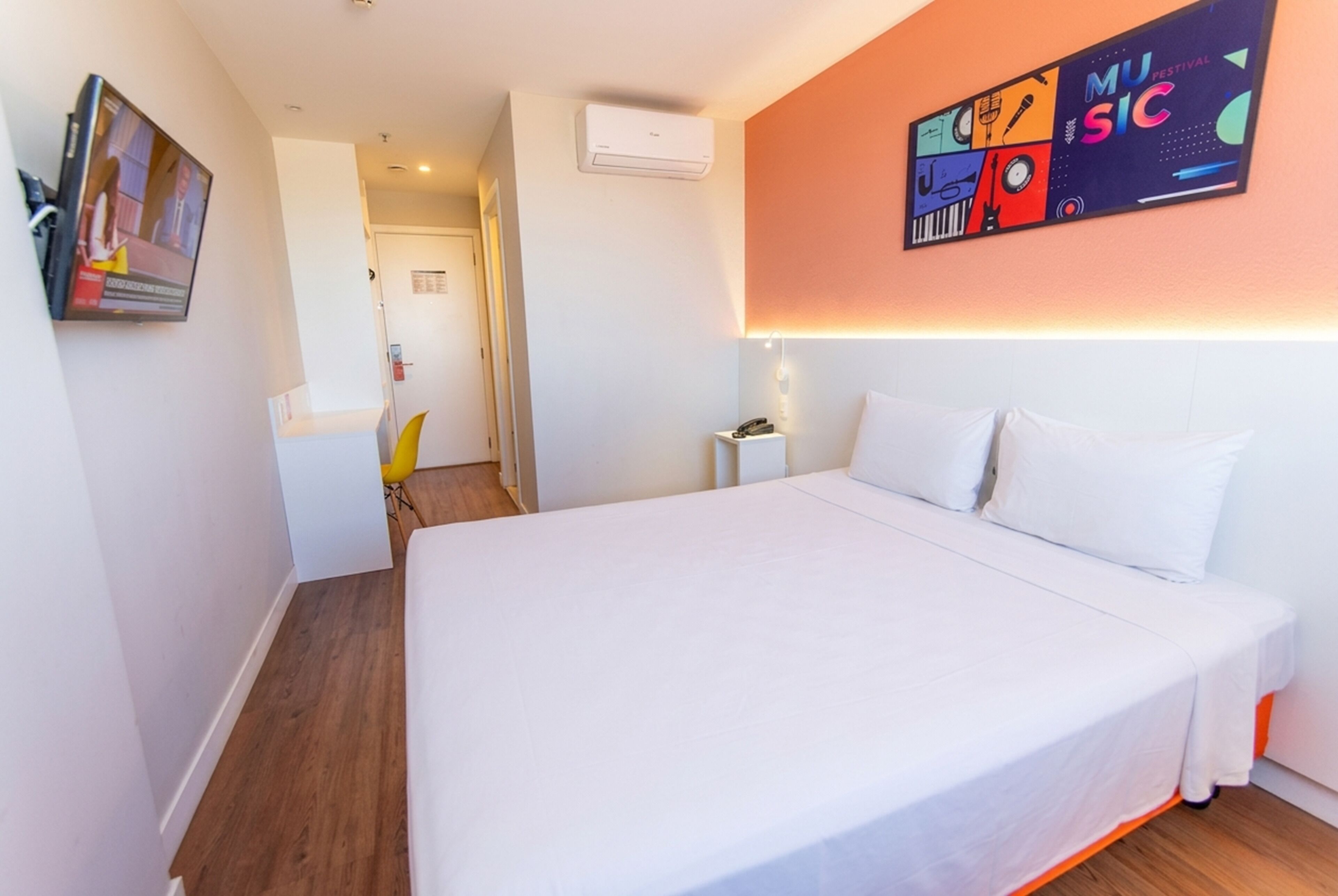 Habitación de hotel con una cama grande, una mesita de noche, aire acondicionado instalado en la pared y un cartel colorido en la pared.