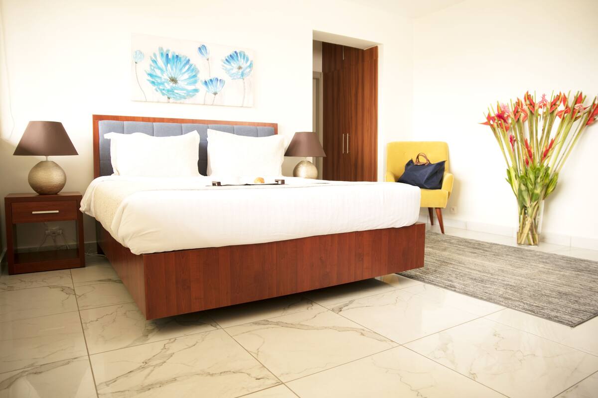 A hotel room with a large bed, two bedside tables with lamps, a wooden wardrobe, a chair, and a vase of flowers.