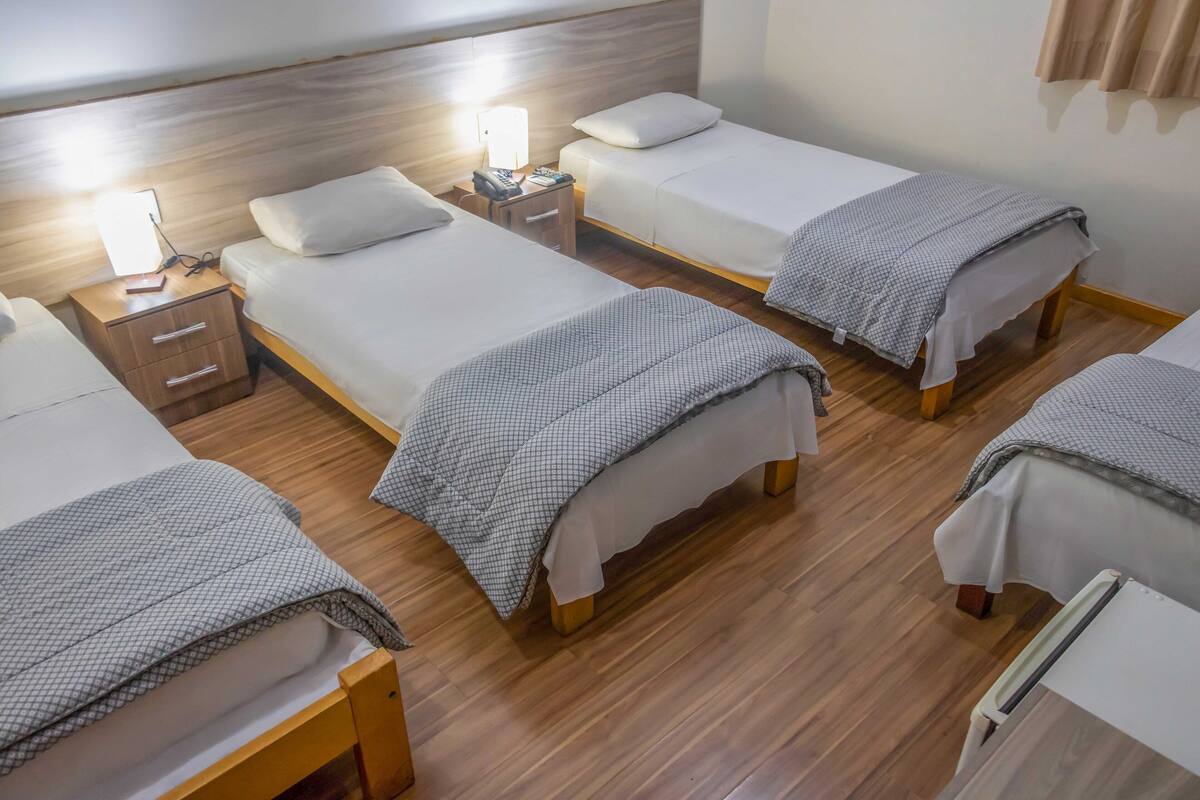 A hotel room with four single beds, wooden flooring, bedside tables with lamps, and a wooden headboard.