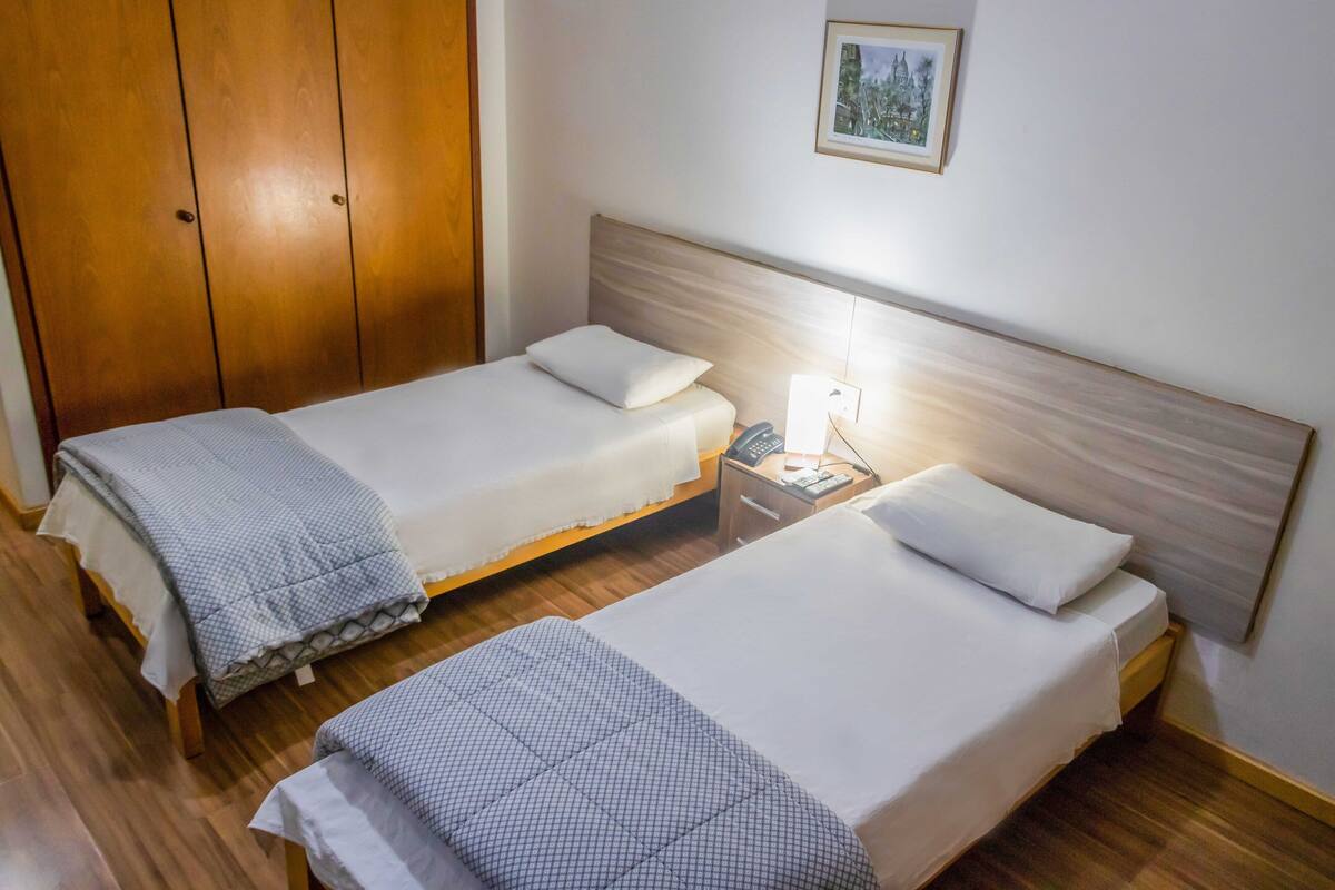 A hotel room with two single beds, a wooden wardrobe, a bedside table with a lamp, and a framed picture on the wall.